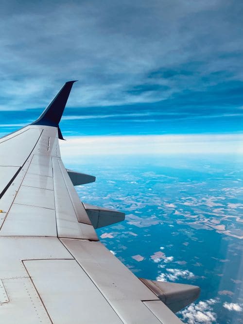 The white wing and blue winglet of an airliner seen from a window, over a cyan sky and farmland far below